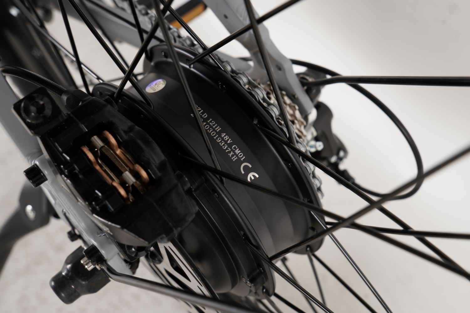 Primer plano de una rueda de bicicleta con radios negros y un motor negro. También son visibles las pastillas de freno y la cadena.