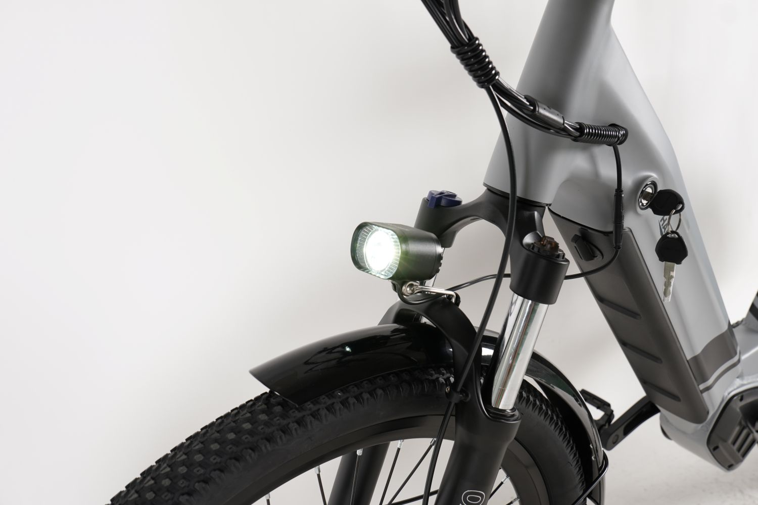 Primer plano de una bicicleta eléctrica gris. Faro brillante, neumático y guardabarros negros, llaves y cableado visibles.