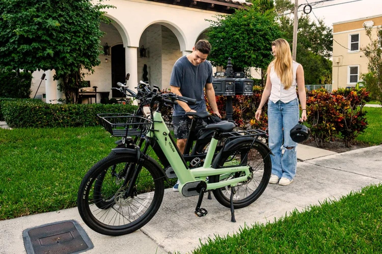 Un hombre y una mujer están cerca de una bicicleta eléctrica verde claro. La mujer sostiene un casco. Al fondo hay una casa y árboles.