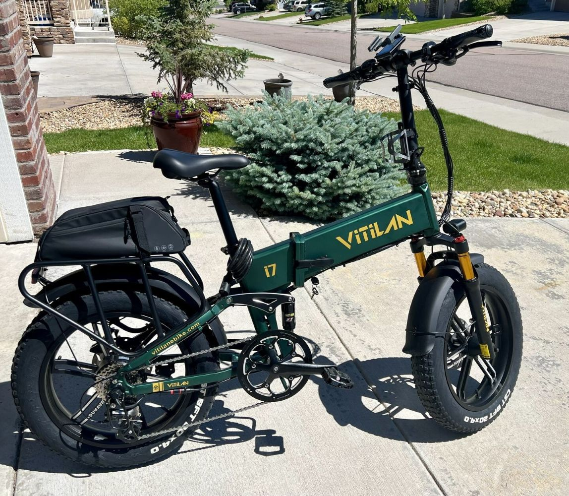 Una bicicleta eléctrica verde Vilian i7 estacionada sobre una superficie de hormigón. La bicicleta tiene una bolsa negra en la parte trasera. El fondo muestra una casa y árboles.