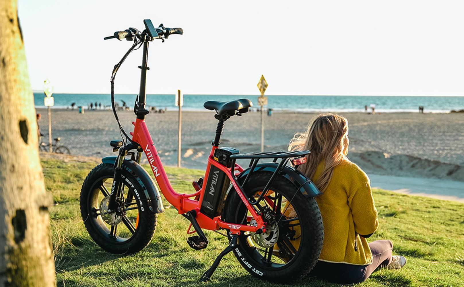 Una e-bike roja Vitilan está sobre la hierba. Una mujer de amarillo está sentada, mirando la playa y el océano. La bici es en un día soleado.