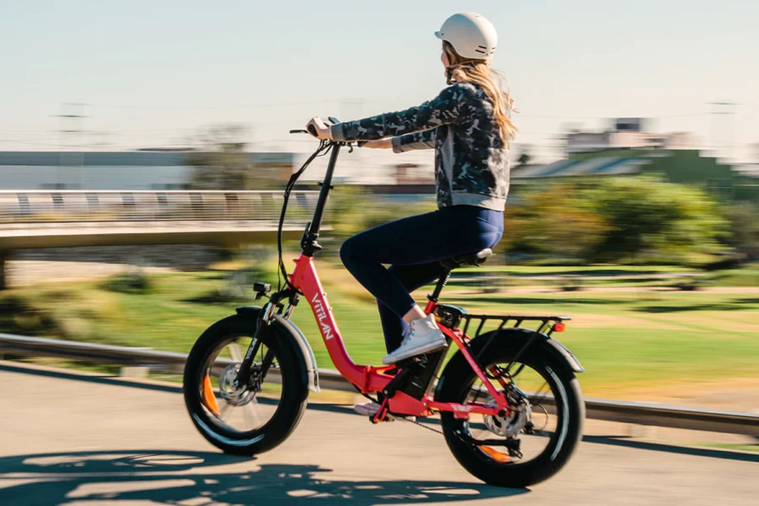 Una donna guida una bici elettrica rossa su una strada, con casco e giacca mimetica, con un ponte sullo sfondo.