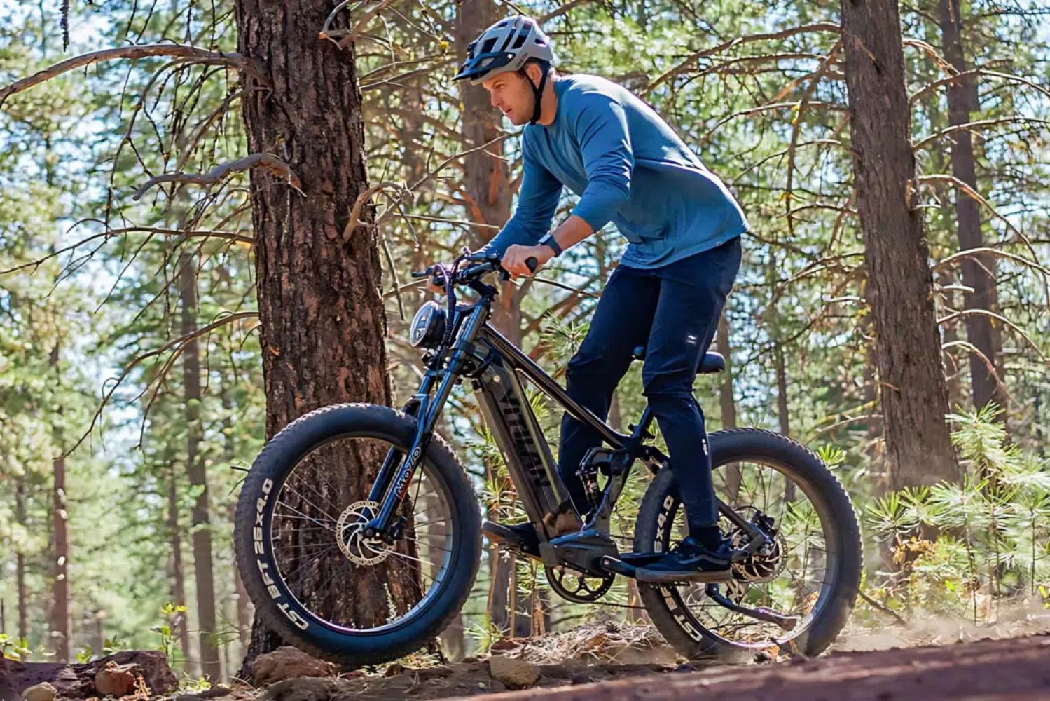 Hombre montando una bicicleta eléctrica negra en un camino de tierra en un bosque. Lleva casco, camisa azul y pantalones oscuros.