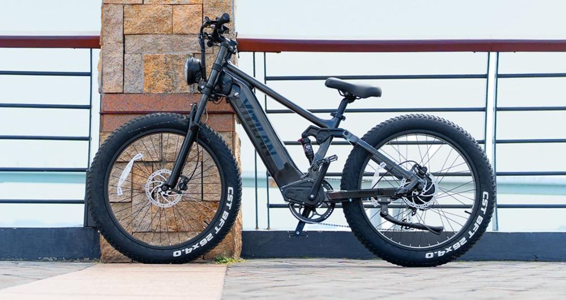 Una bicicleta eléctrica negra está estacionada. Tiene neumáticos anchos, un faro y está al lado de un pilar de piedra y una barandilla de metal.