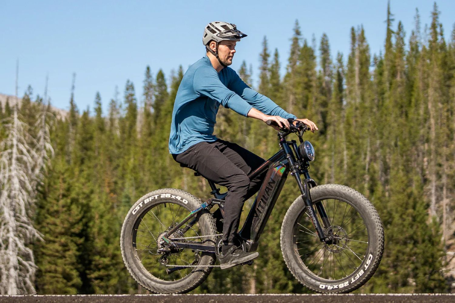 Un hombre monta una bicicleta eléctrica negra en una carretera. Fondo de bosque y cielo azul claro. Lleva casco y camisa azul.