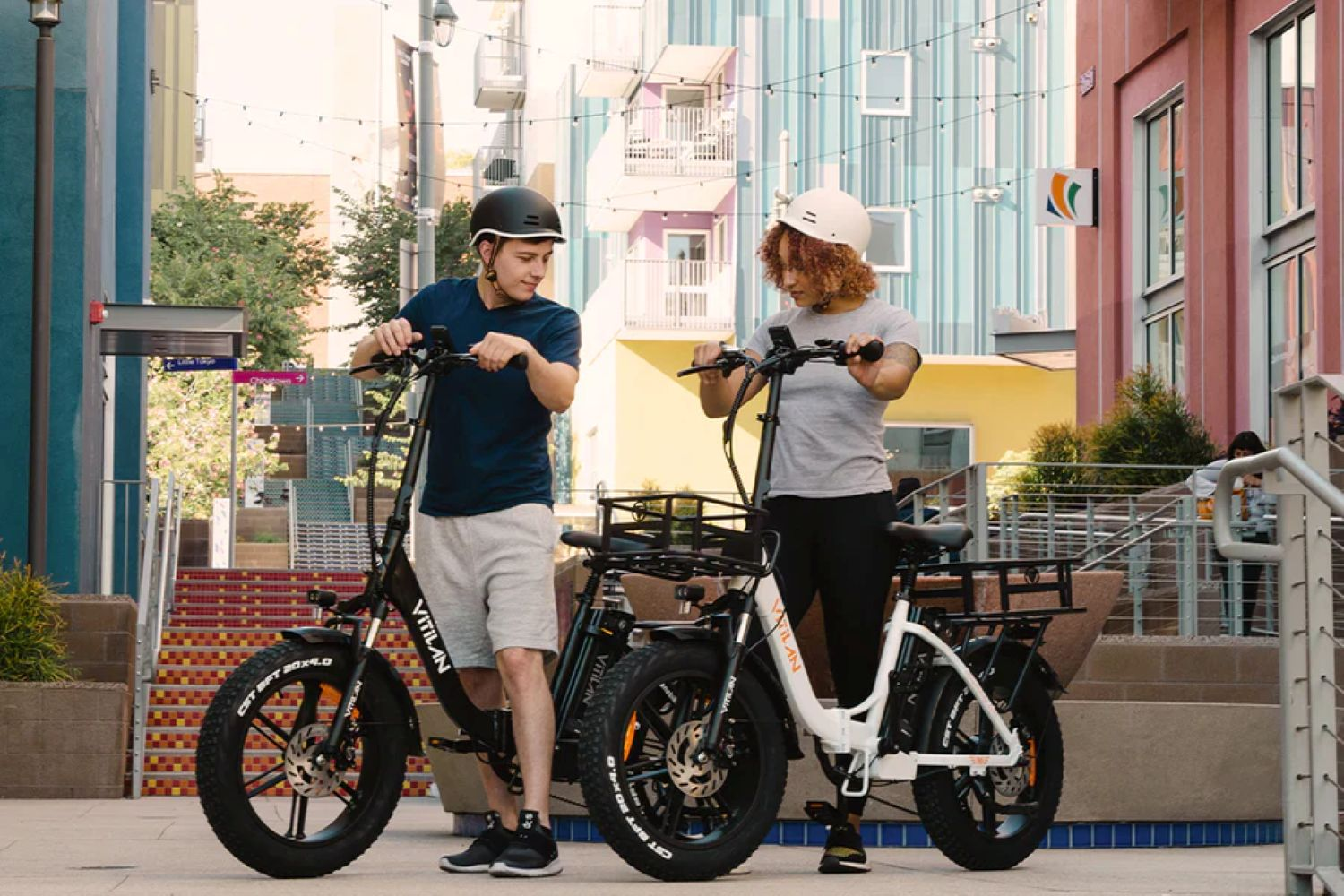 Dos personas con cascos junto a e-bikes. Edificios los rodean. Una bicicleta es negra, la otra blanca.