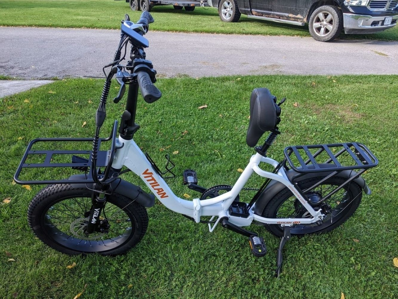 Bicicleta eléctrica Vilitan blanca sobre hierba verde. Tiene portaequipajes delanteros y traseros. Una camioneta negra y un remolque están en el fondo.