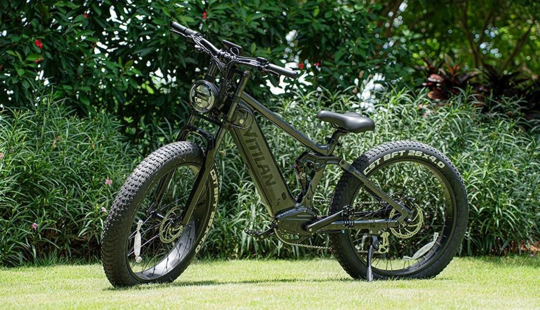Una bicicleta eléctrica negra está estacionada en la hierba. Arbustos y árboles forman el fondo. La bicicleta tiene llantas anchas y un faro.