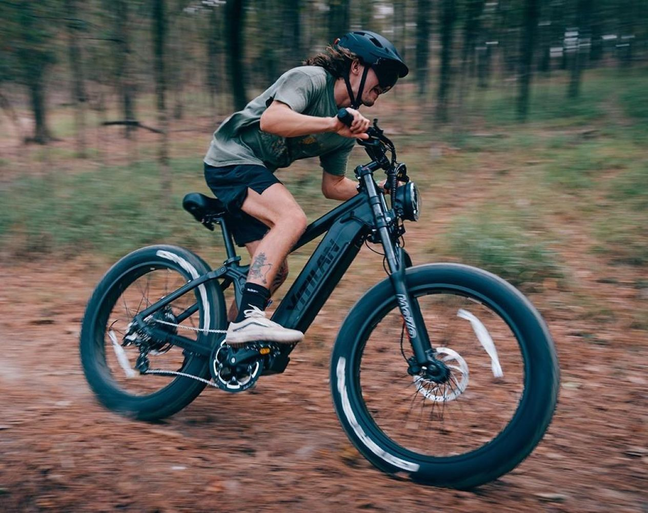 Hombre montando una bicicleta eléctrica negra en un camino de tierra en un bosque. Lleva casco, pantalones cortos y camiseta. Los árboles se difuminan en el fondo.