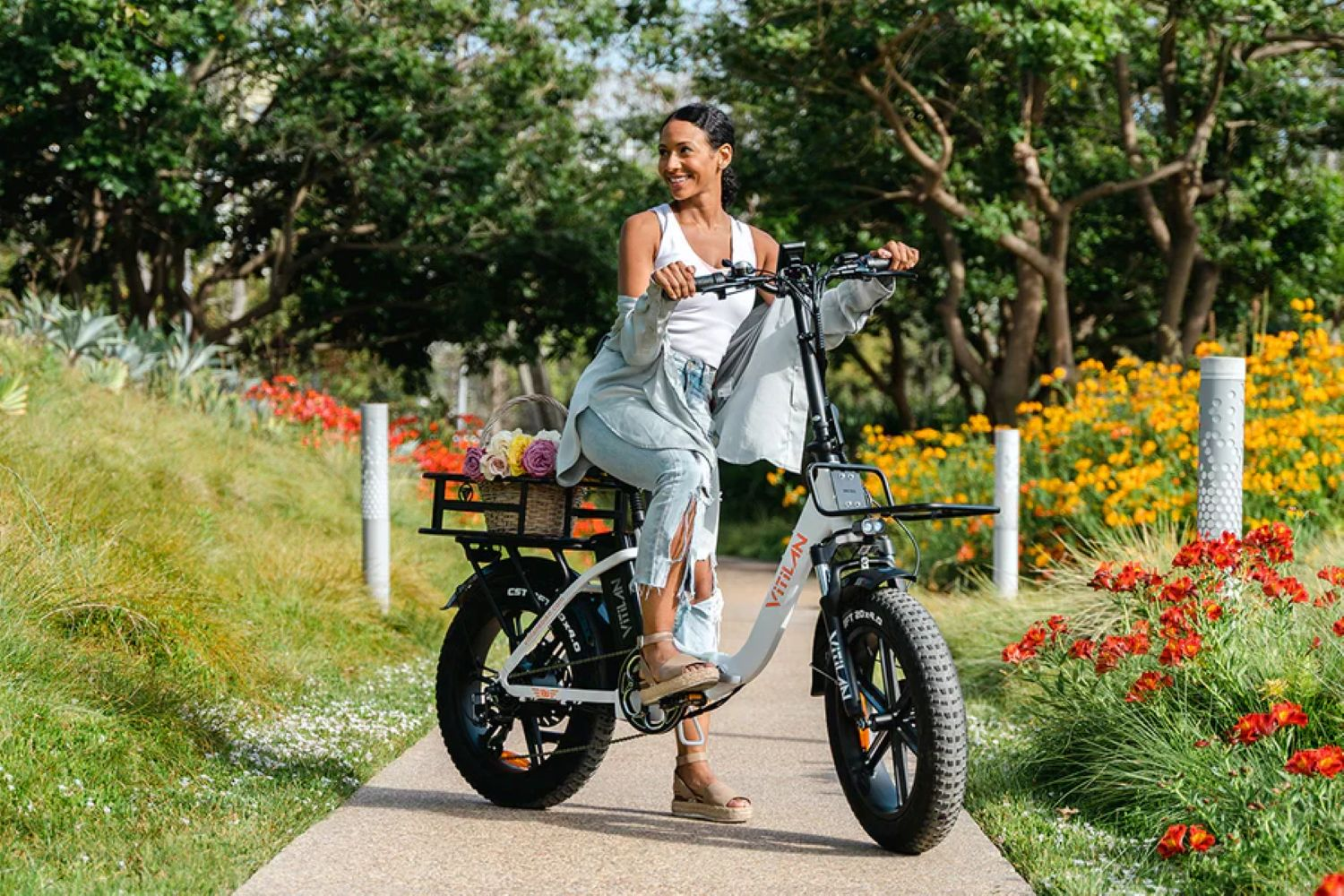 Una mujer en una bicicleta eléctrica sonríe. Tiene una cesta de flores. Flores y vegetación rodean la bicicleta en un camino.