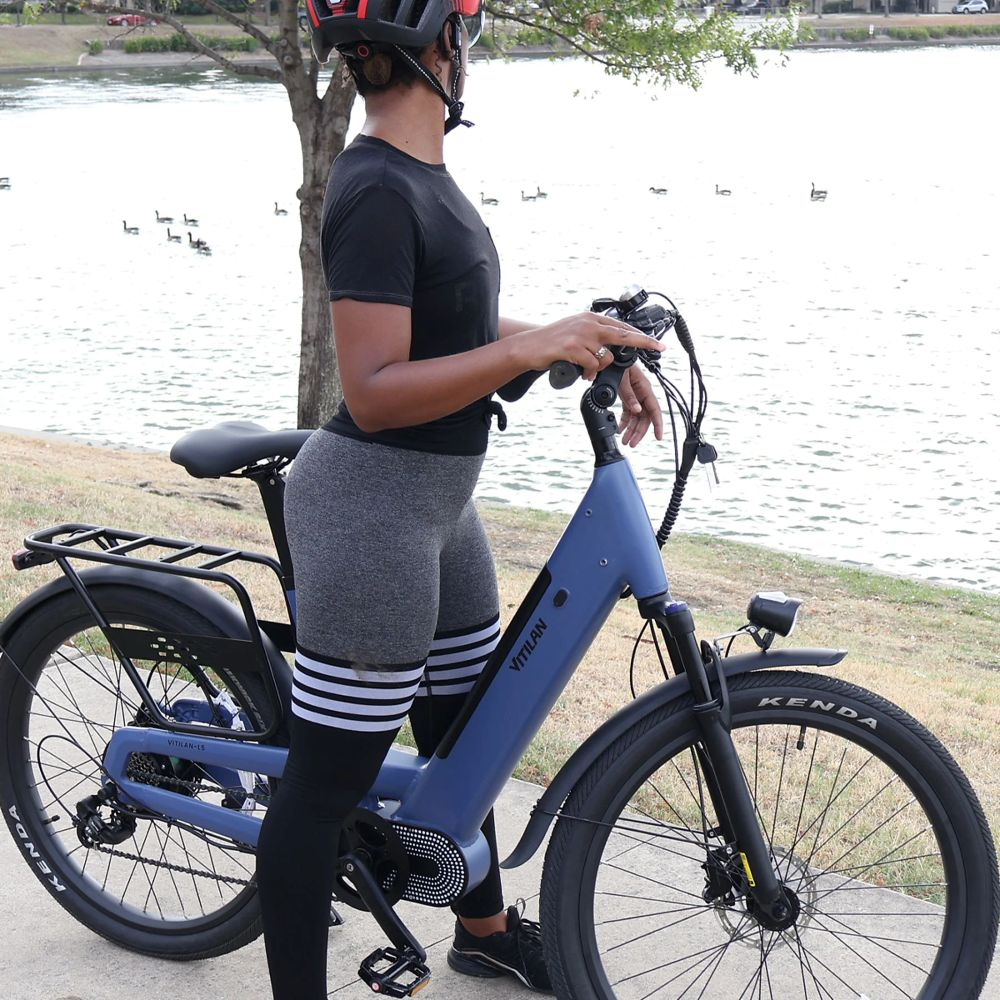 Una mujer con casco está junto a una bicicleta eléctrica azul cerca de un lago. Lleva camisa negra, leggings grises. Patos nadan.
