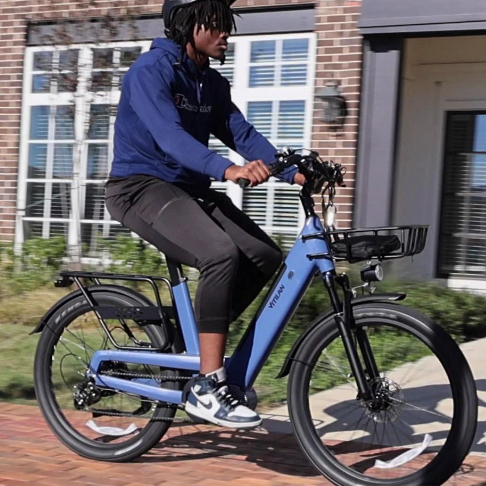 Una persona monta una bicicleta eléctrica azul en un camino de ladrillos. Viste una sudadera con capucha azul, pantalones negros y un casco. Un edificio está al fondo.