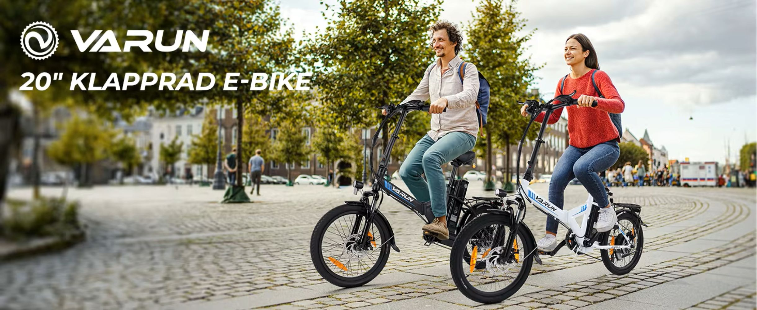 Dos personas montan bicicletas eléctricas en una calle adoquinada. Árboles y edificios al fondo, una pancarta dice 'VARUN E-BIKE'.