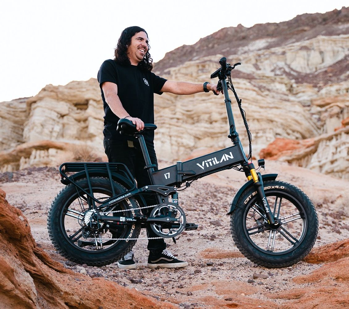 Un hombre sonríe junto a una e-bike Vitilan negra. Viste de negro. El fondo muestra formaciones rocosas desérticas.