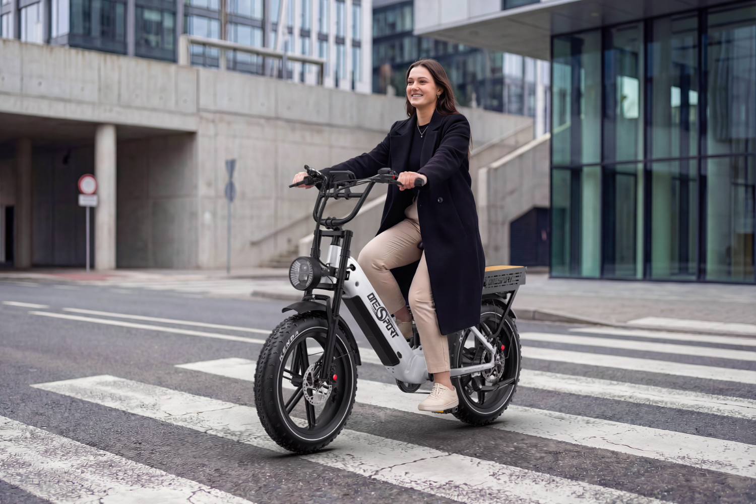 Una mujer monta en una bicicleta eléctrica negra y blanca por un paso de peatones. Lleva un abrigo negro y pantalones color beige.