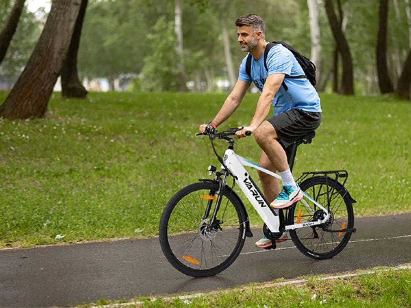 Un hombre monta una bicicleta eléctrica blanca y negra en un camino. Lleva camisa azul y pantalones cortos negros. Hay hierba verde y árboles de fondo.