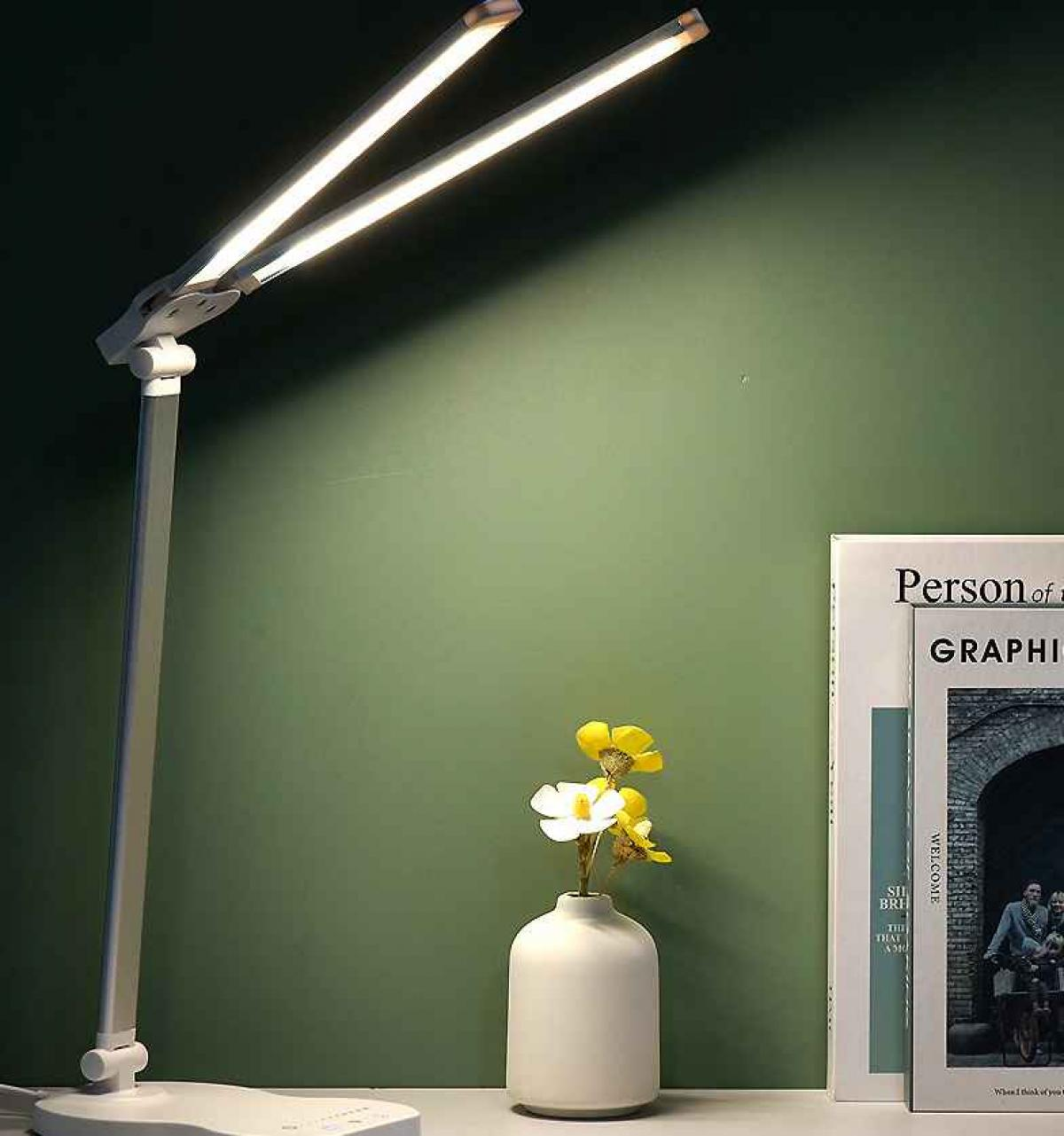 Eine weiße Schreibtischlampe mit zwei Lichtleisten beleuchtet eine Blumenvase und ein Buch vor einer grünen Wand.