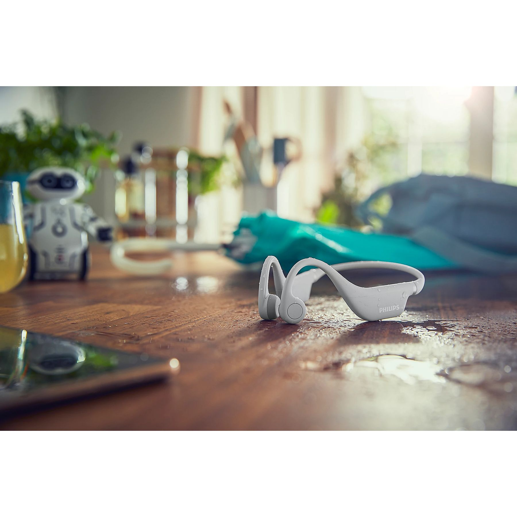 Białe słuchawki Philips na mokrym drewnianym stole. Obecny jest również robot-zabawka, parasol i telefon.