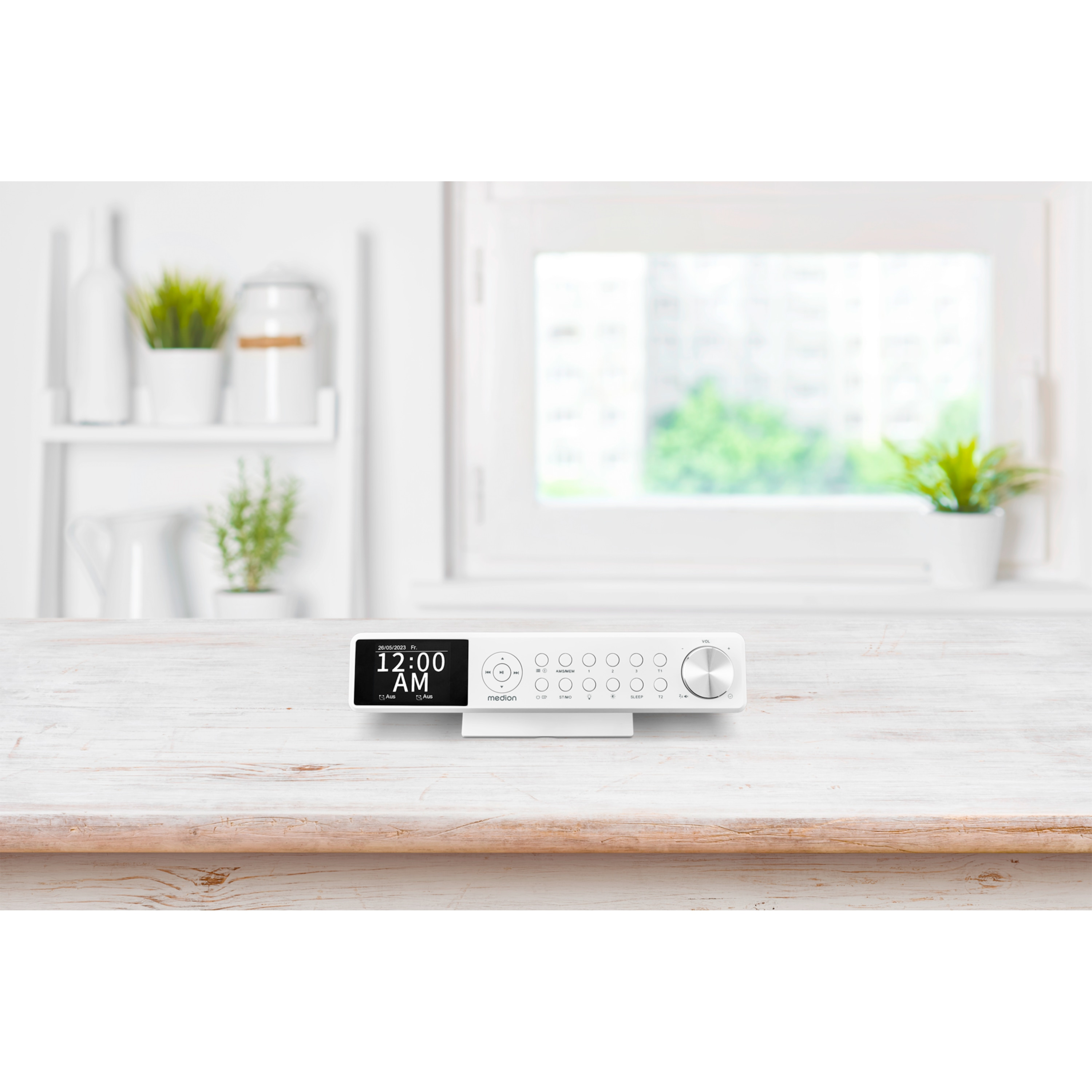 A white digital clock radio displaying 12:00 AM sits on a wooden table. A window and plants are in the background.