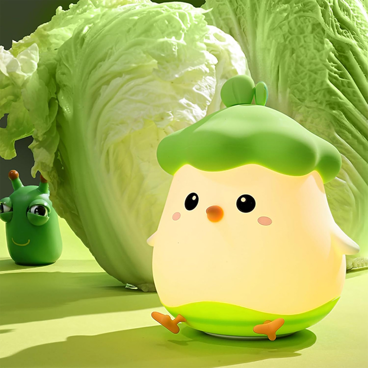 Eine cartoonartige, kükenförmige Lampe mit grünem Hut steht vor Kohl. Ein grünes Raupenspielzeug befindet sich links. Die Szene ist hell.