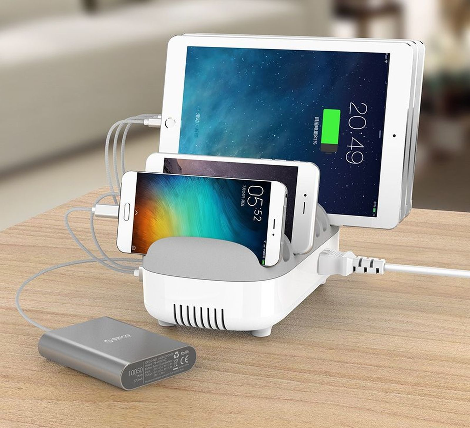 Eine weiße Ladestation hält ein Tablet und zwei Telefone. Eine silberne Powerbank liegt auf der Holzoberfläche.