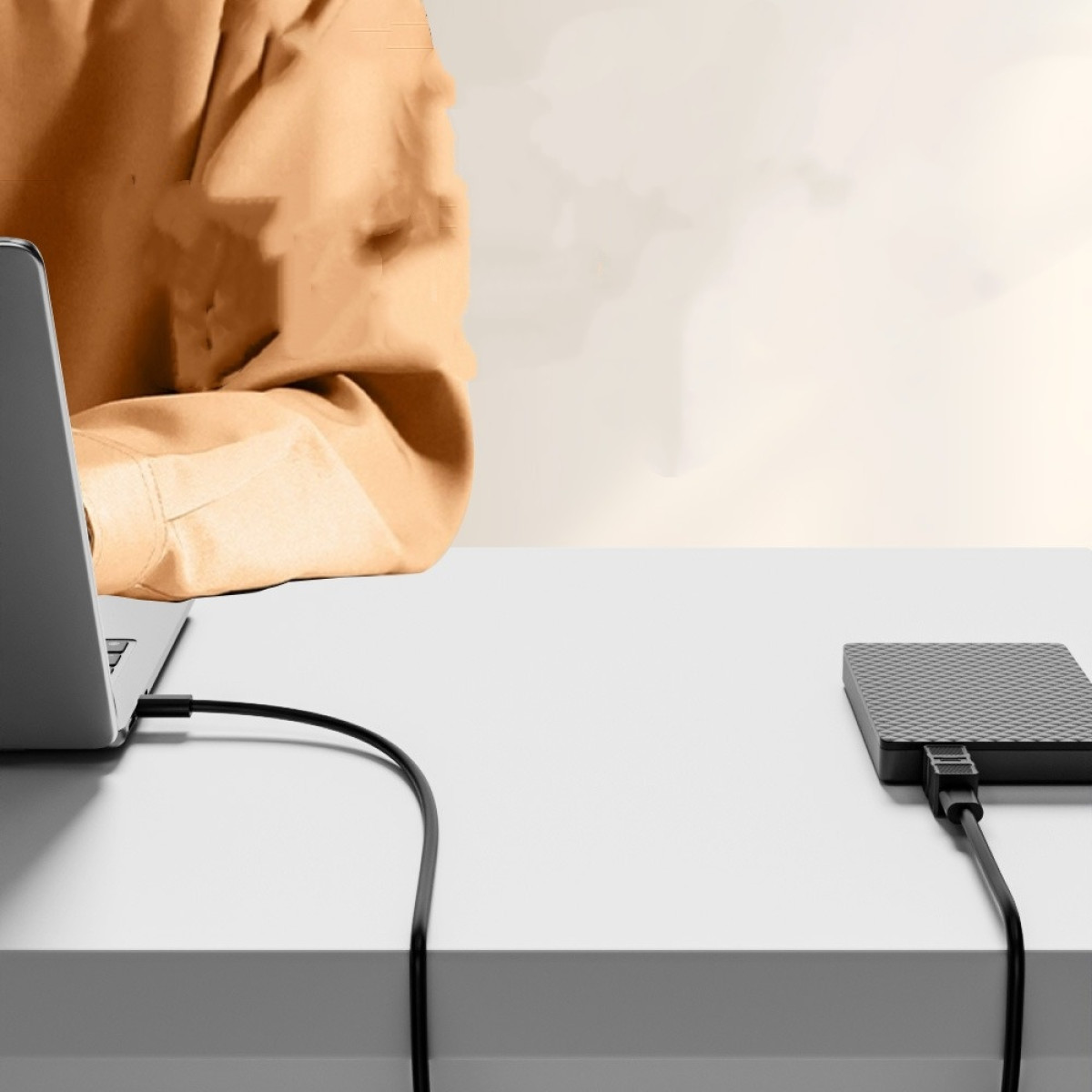 Laptop ist mit einer Festplatte auf einem weißen Schreibtisch verbunden. Eine Person in einem orangefarbenen Hemd ist teilweise sichtbar.