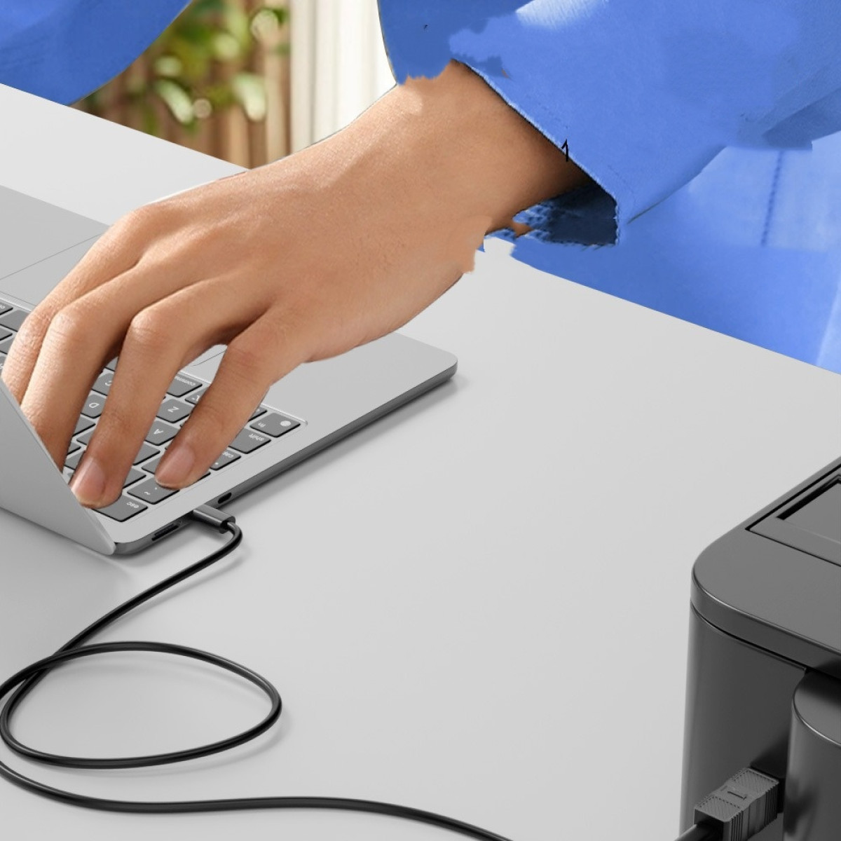 Eine Hand berührt eine Laptop-Tastatur, die mit einem schwarzen Kabel verbunden ist. Ein blauer Ärmel ist sichtbar. Rechts ist ein schwarzes Gerät.