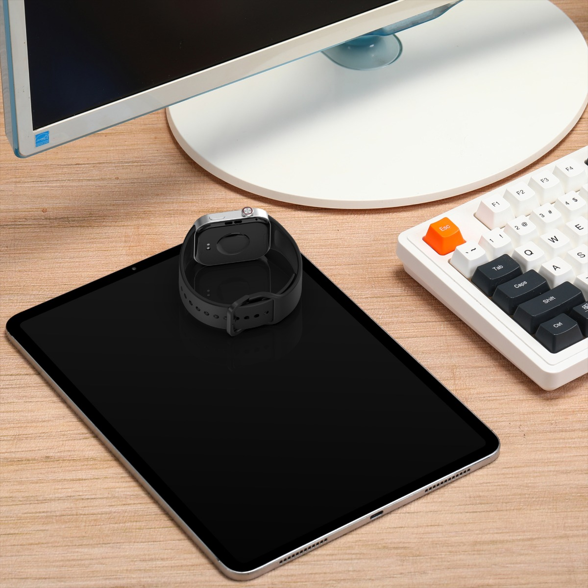Eine Smartwatch liegt auf einem Tablet. Eine Tastatur und ein Computerbildschirm befinden sich im Hintergrund, alles auf einer Holzoberfläche.