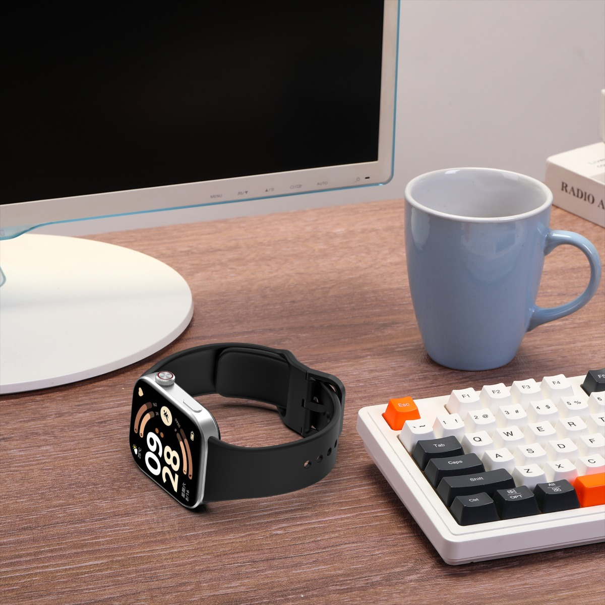 Eine Smartwatch mit schwarzem Armband liegt auf einem Holzschreibtisch neben einer Tastatur, einer Tasse und einem Computermonitor.