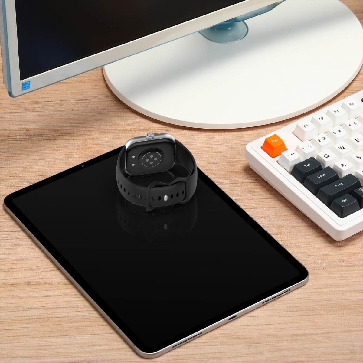 Eine Smartwatch liegt auf einem Tablet. Eine Tastatur und ein Monitor befinden sich im Hintergrund, alles auf Holz.