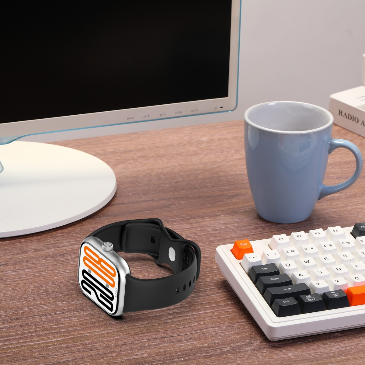 Eine Smartwatch mit schwarzem Armband liegt auf einem Holzschreibtisch. Daneben befinden sich eine Tastatur, eine blaue Tasse und ein Computermonitor.