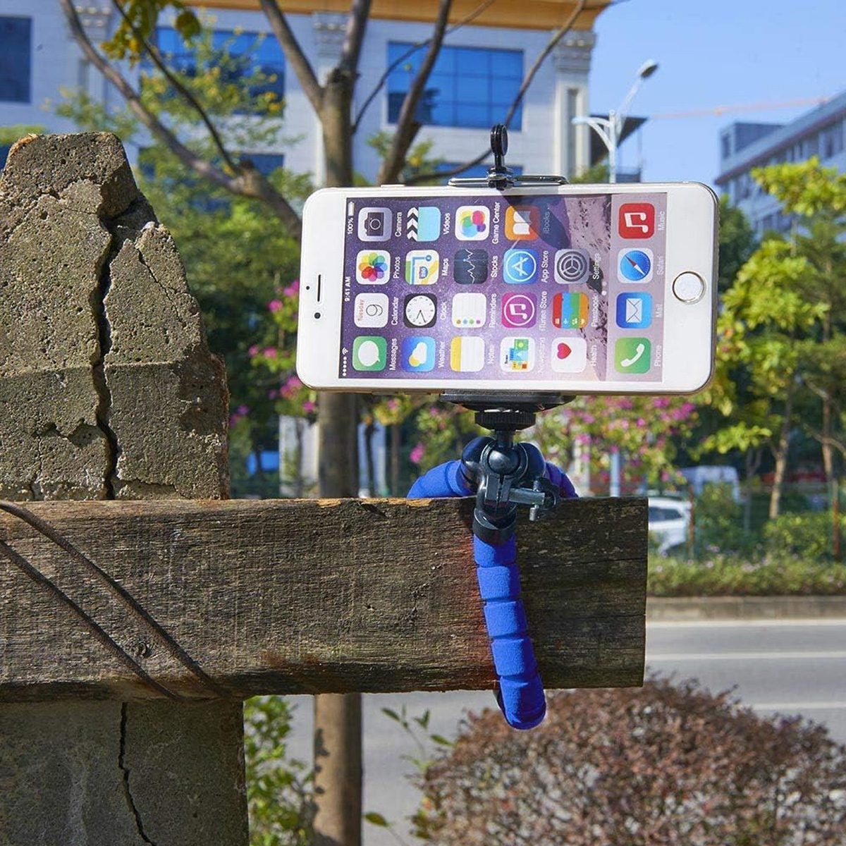 Ein Smartphone ist auf einem blauen Stativ montiert, das an einem Holzbalken befestigt ist. Der Hintergrund zeigt Bäume, Gebäude und eine Straße.