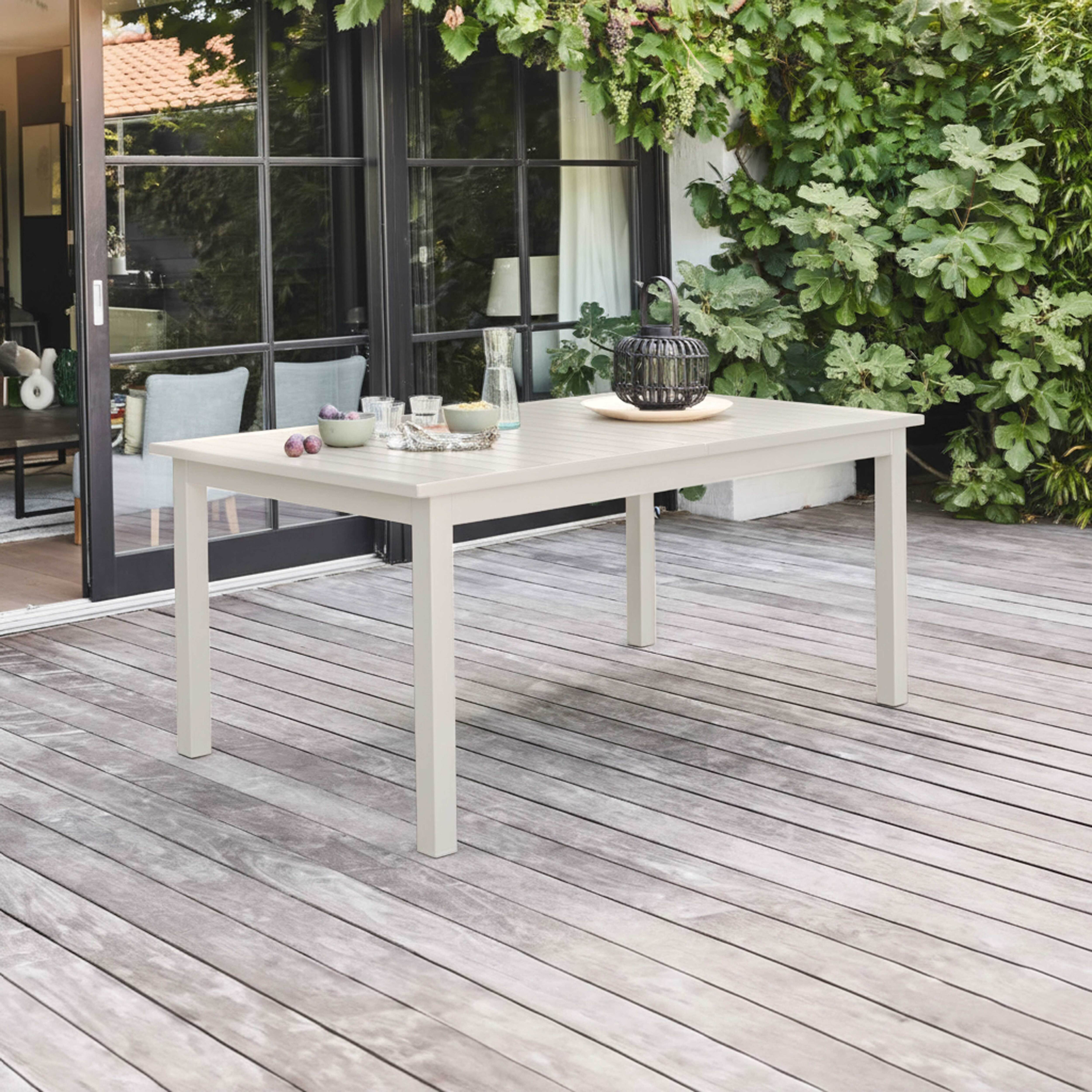 Ein weißer Tisch mit Gegenständen steht auf einer Holzterrasse. Grüne Pflanzen und eine Glastür im Hintergrund.