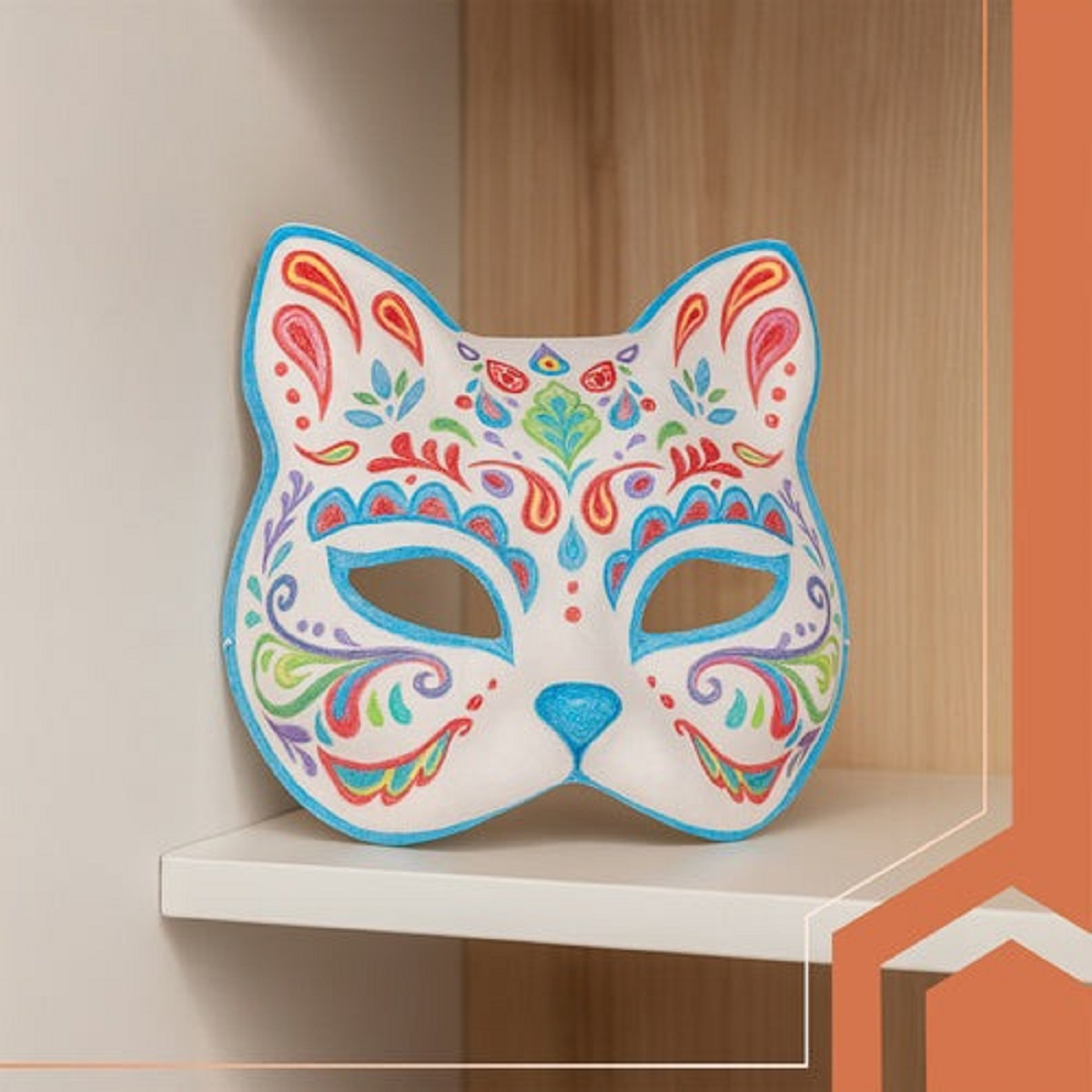 Una máscara de gato decorativa con contorno azul y patrones florales coloridos sobre fondo blanco, colocada en un estante blanco.