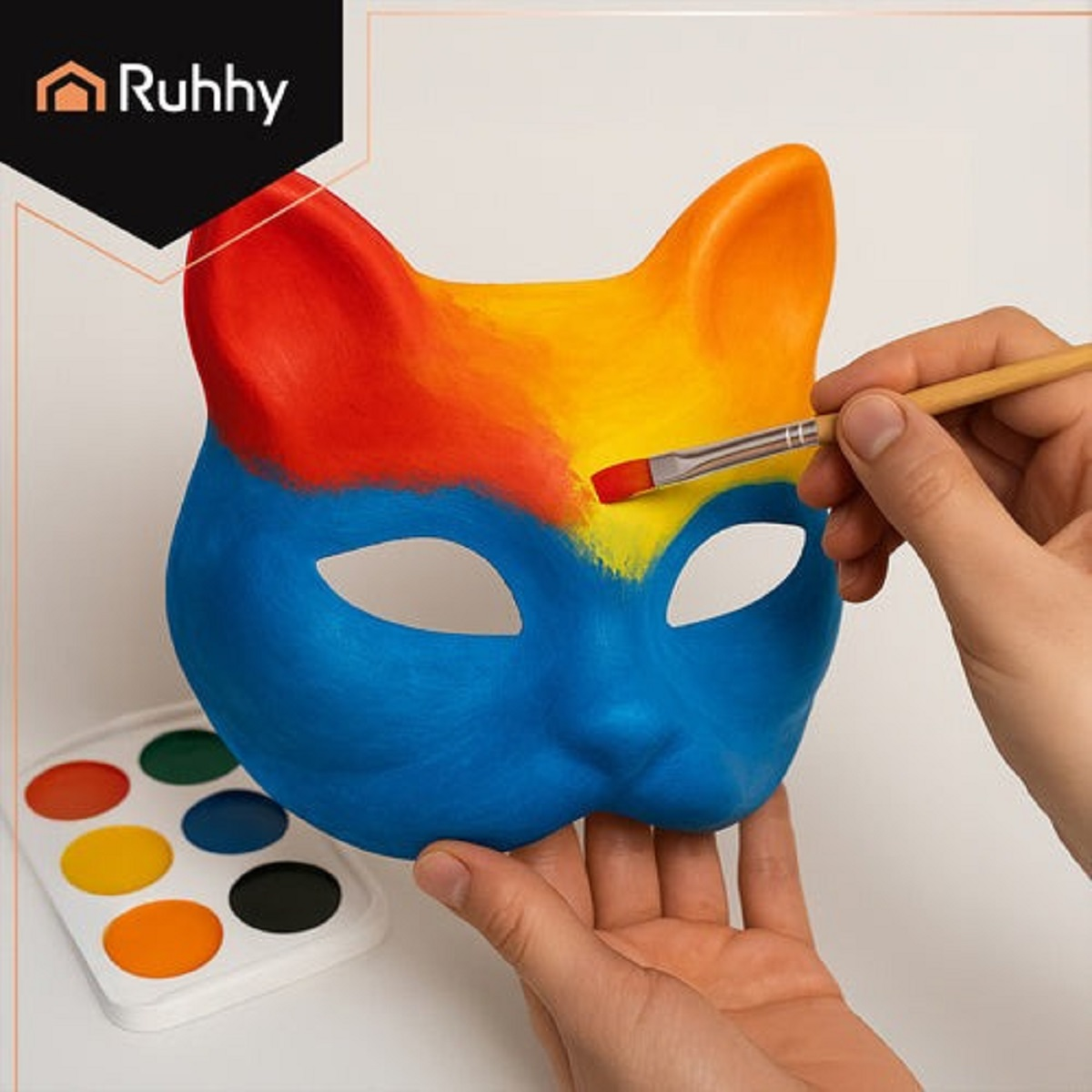 Una máscara de gato siendo pintada con un pincel, con secciones azules, amarillas y rojas. Una paleta de colores está al lado.