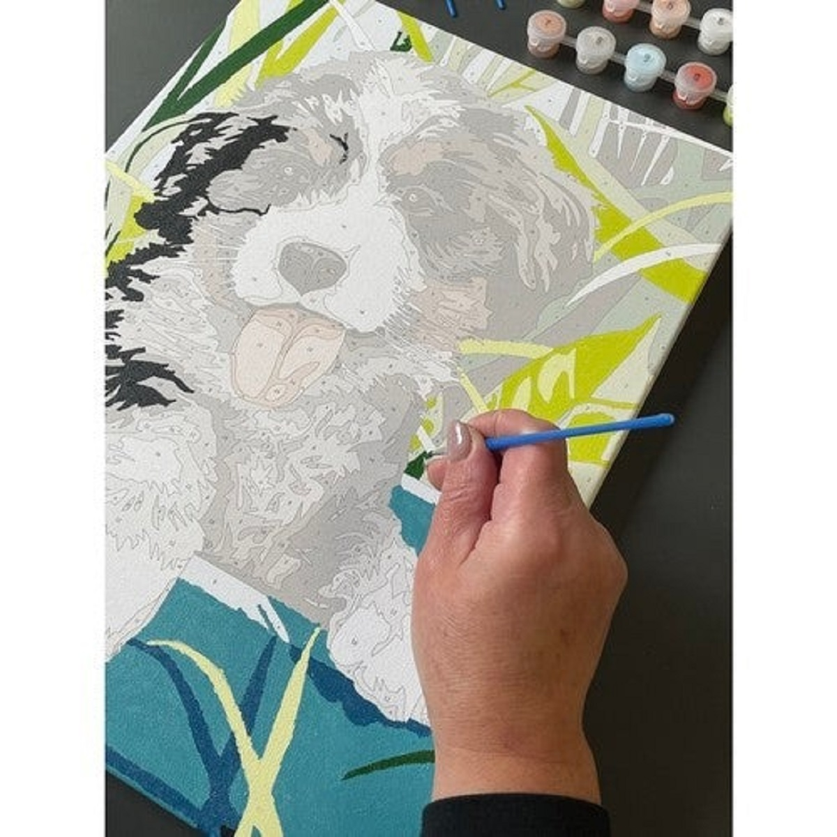 Una mano con un pincel azul pinta un perro en un lienzo. El perro es principalmente gris y blanco, con la lengua fuera. Pinturas en el fondo.