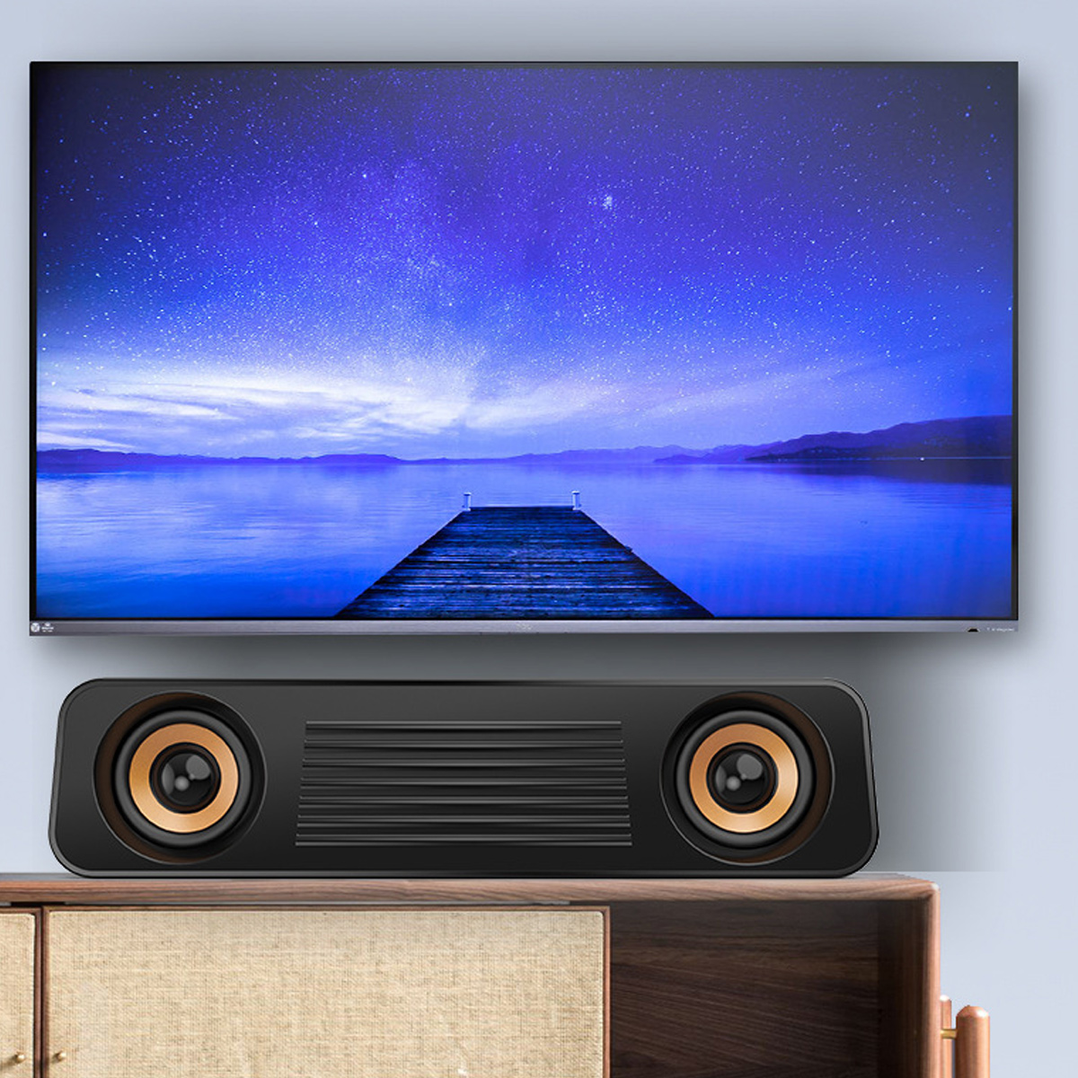 Ein Fernseher zeigt eine nächtliche Seeszene mit Sternen. Unterhalb befindet sich ein schwarzer Lautsprecher mit goldenen Akzenten. Der Lautsprecher steht auf einem Holzschrank.