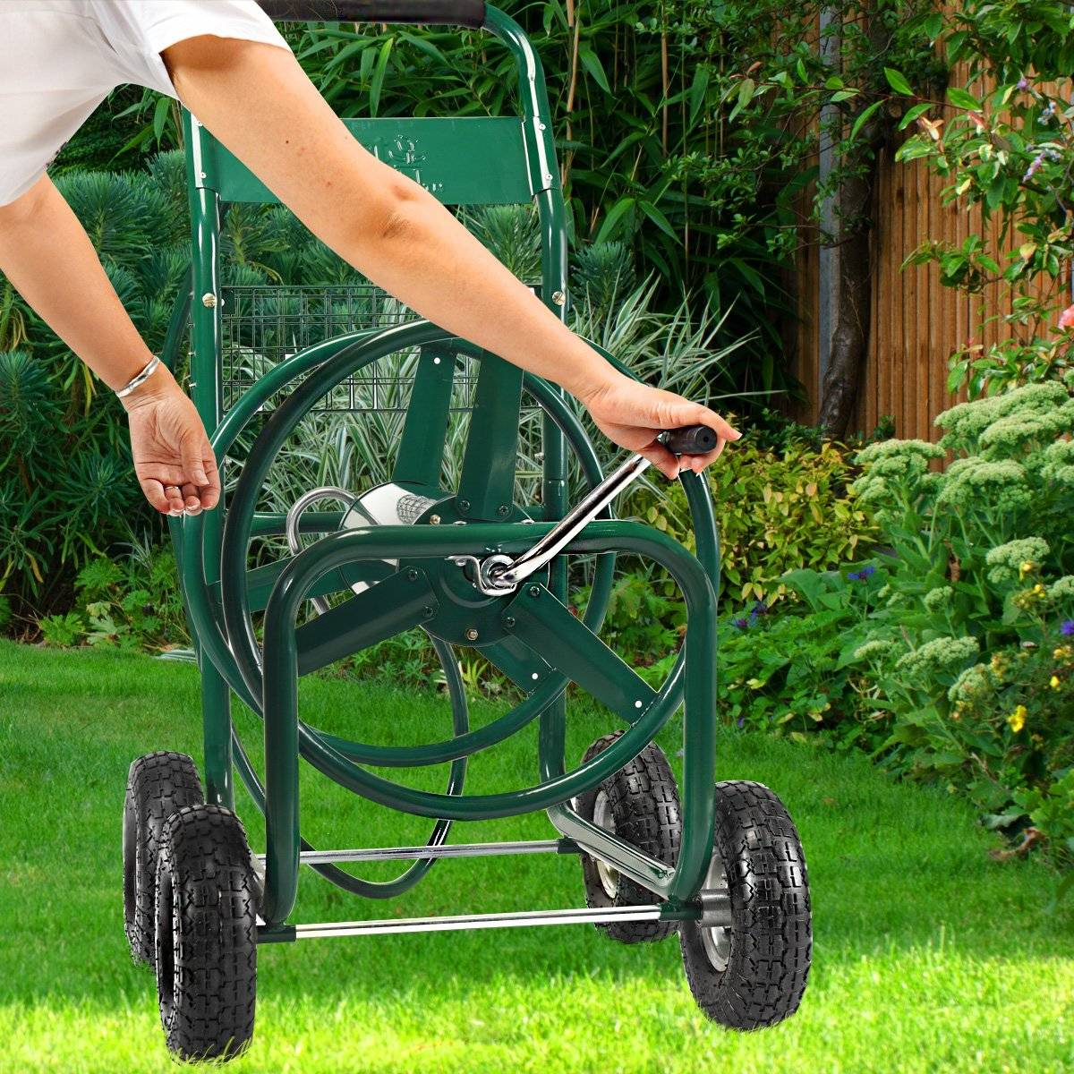 Una persona usa un carrete de manguera verde sobre ruedas en un jardín. La persona está girando la manivela. Césped verde y plantas en el fondo.
