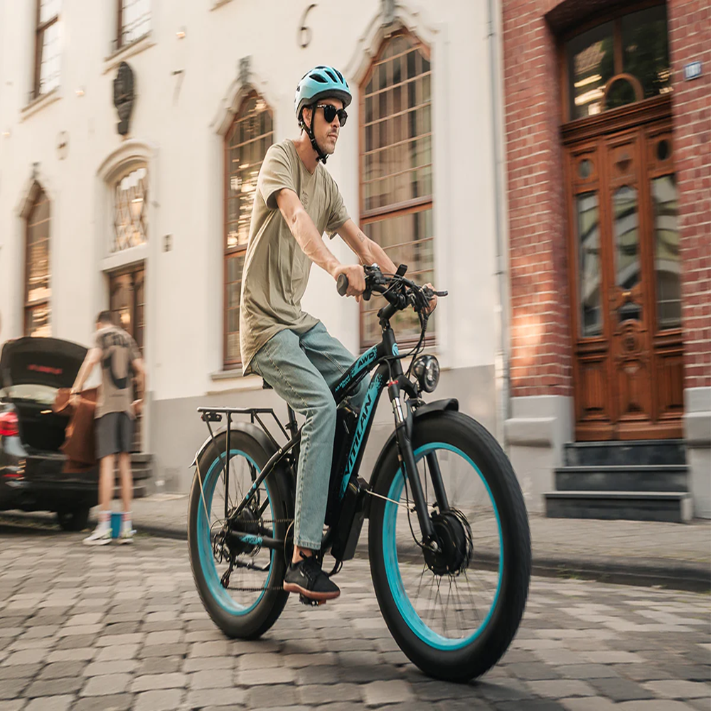 Un hombre monta en bicicleta eléctrica negra y azul sobre adoquines. Lleva casco y gafas de sol. Un coche y edificios en el fondo.