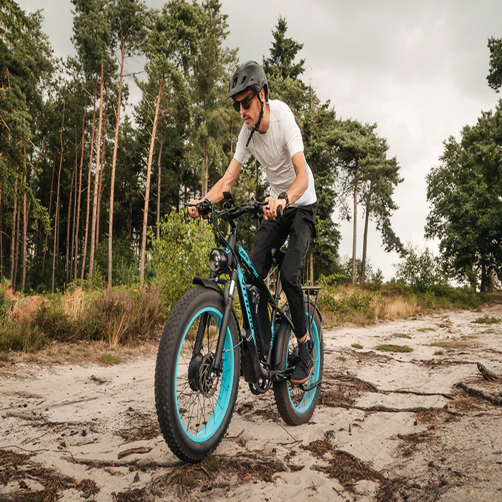 Un hombre monta en bicicleta eléctrica por un camino de tierra. La bicicleta tiene detalles azules. Árboles al fondo. Cielo nublado.