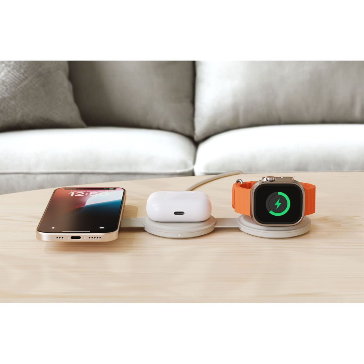 Ein Telefon, AirPods und eine Uhr befinden sich auf einer Ladestation. Die Uhr zeigt ein grünes Ladesymbol. Der Hintergrund zeigt ein Sofa.
