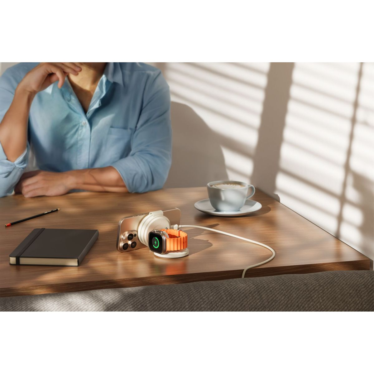 Eine Person sitzt an einem Schreibtisch. Ein Telefon und eine Uhr laden an einem Ständer. Eine Kaffeetasse und ein Notizbuch liegen ebenfalls auf dem Schreibtisch.