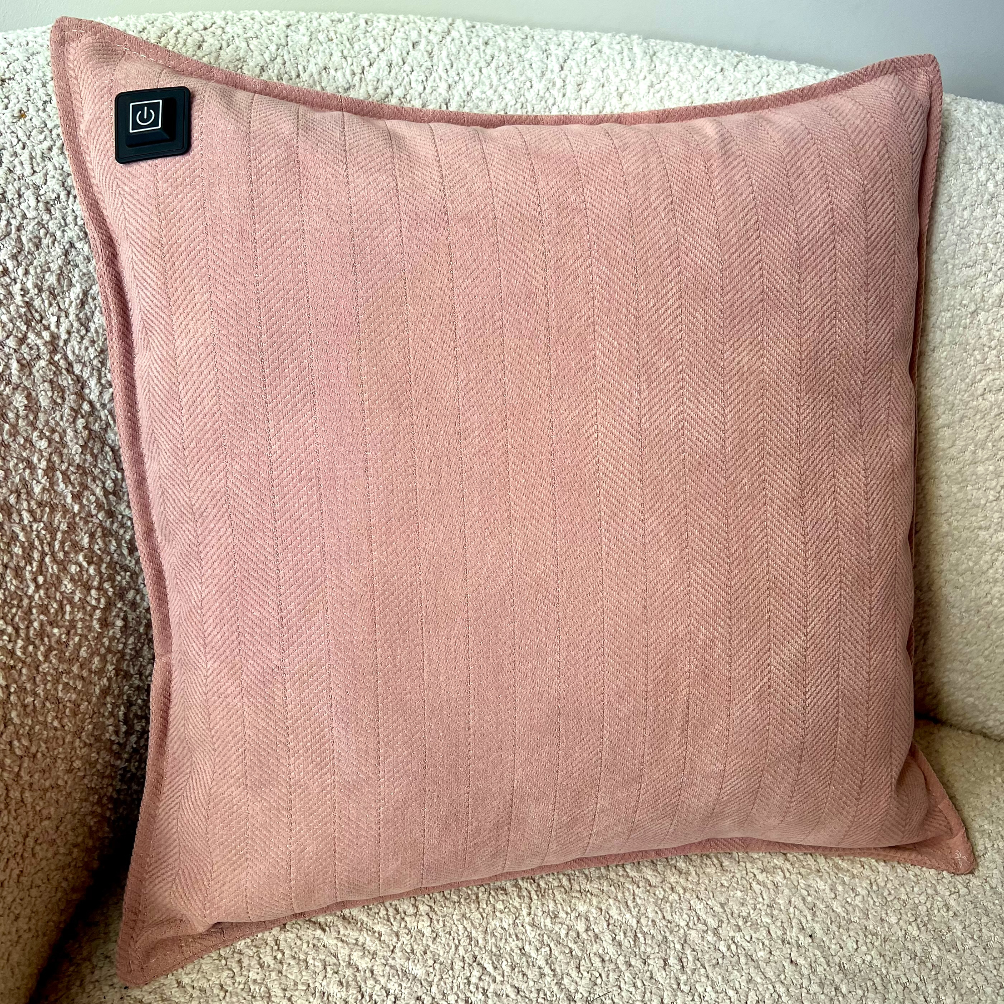 Ein rosa Kissen mit Fischgrätenmuster liegt auf einer strukturierten weißen Oberfläche. Es hat einen Power-Knopf.