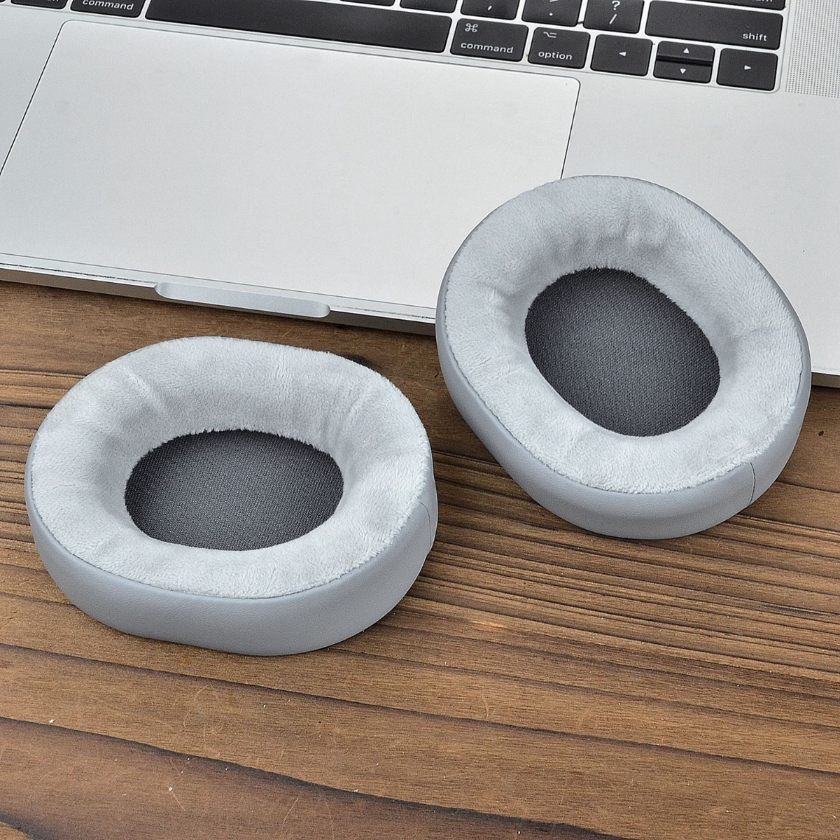 Dos almohadillas grises para auriculares sobre una superficie de madera, cerca de una computadora portátil. Las almohadillas tienen un interior blanco suave y un exterior gris.