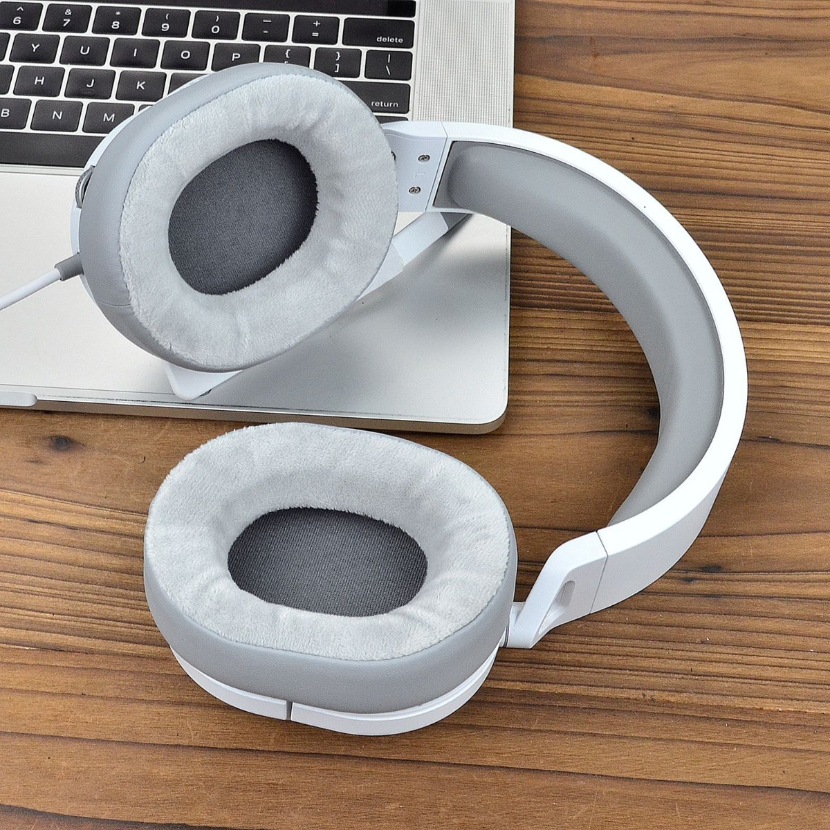 Auriculares con almohadillas grises y marco blanco sobre un teclado de portátil y una mesa de madera. Las almohadillas son suaves.
