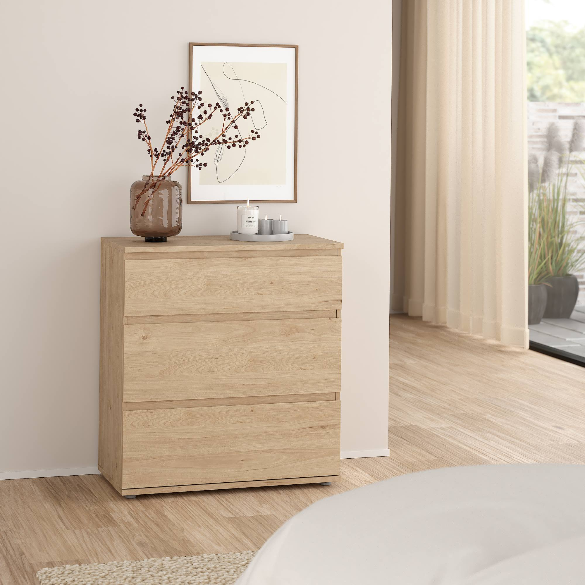 Cómoda de madera con tres cajones, jarrón con ramas, arte enmarcado, velas y ventana con cortinas.