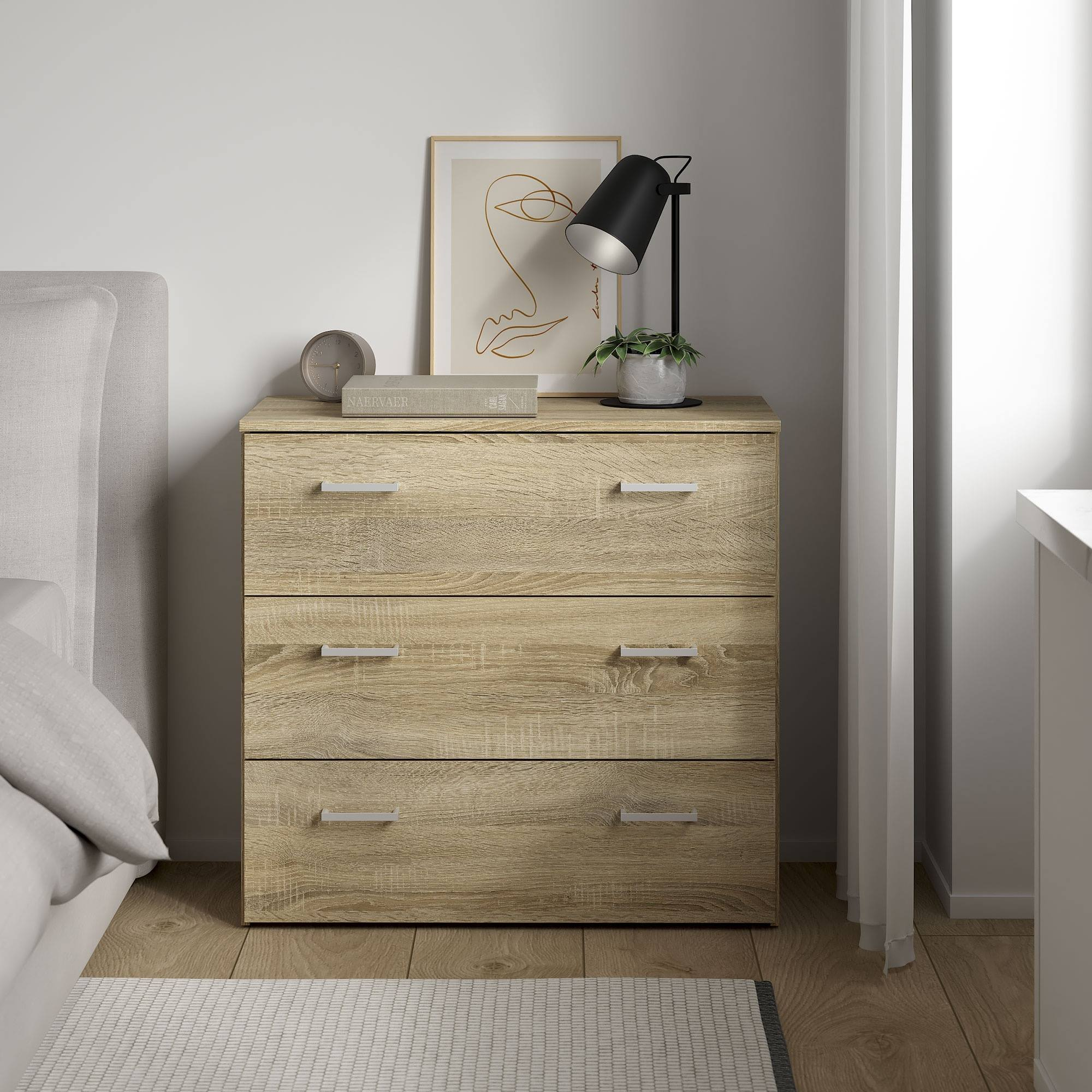 Mesa de noche de madera con tres cajones, lámpara, obra de arte y reloj. Cama a la izquierda, cortinas a la derecha.