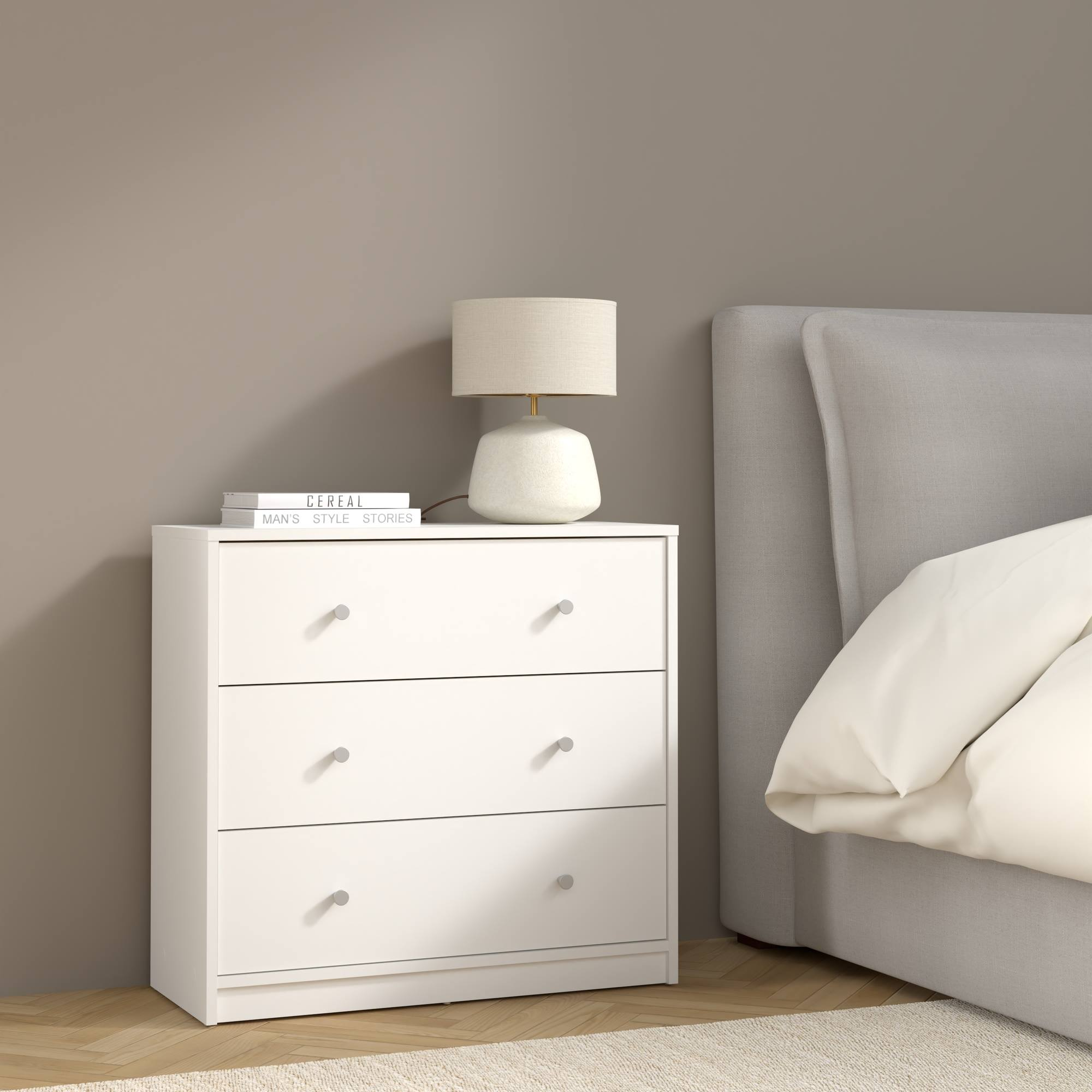 Cómoda blanca con tres cajones, una lámpara y libros. Una cama está en el lado derecho de la imagen.