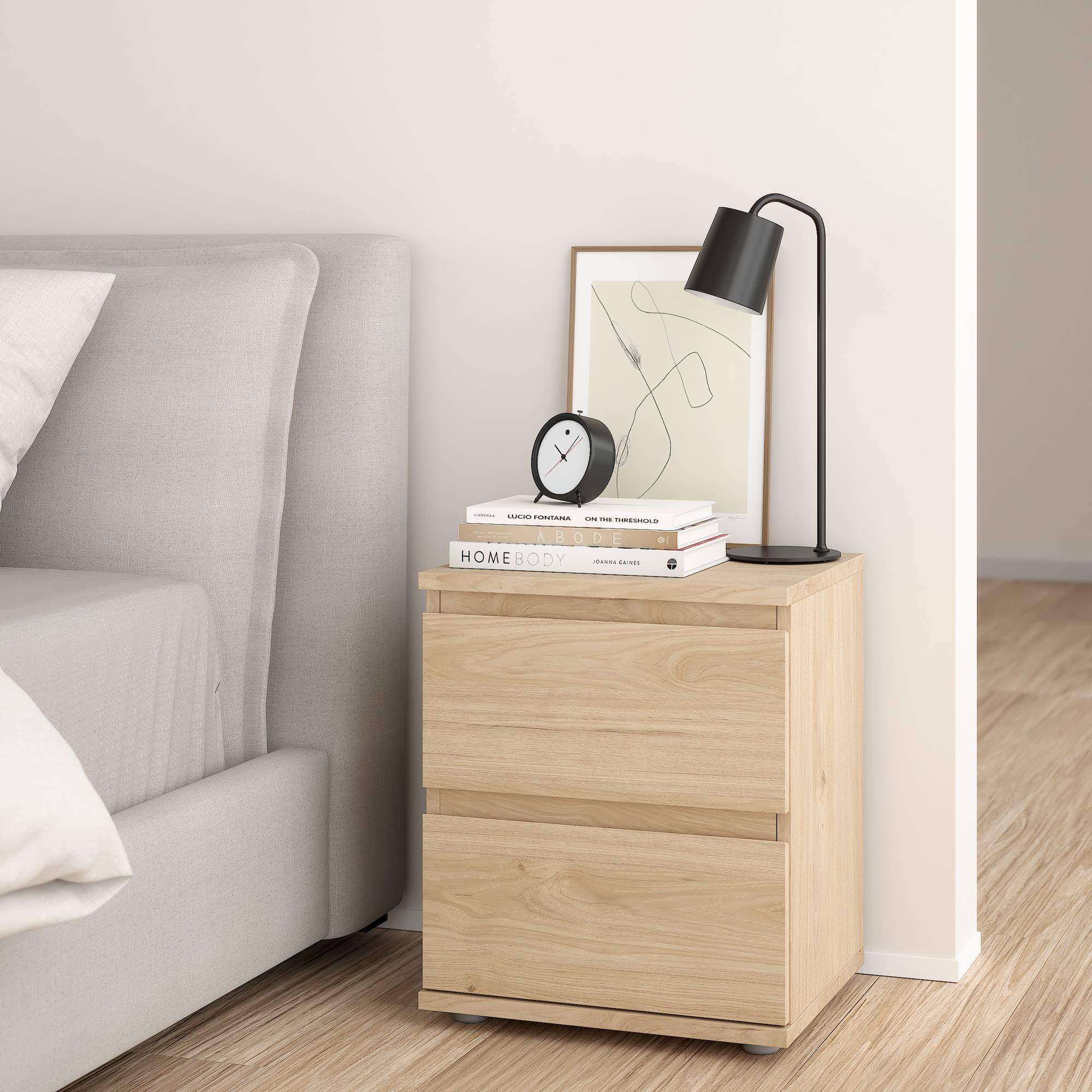 Una mesita de noche con lámpara, reloj y libros. A la izquierda hay una cama. El suelo es de madera.
