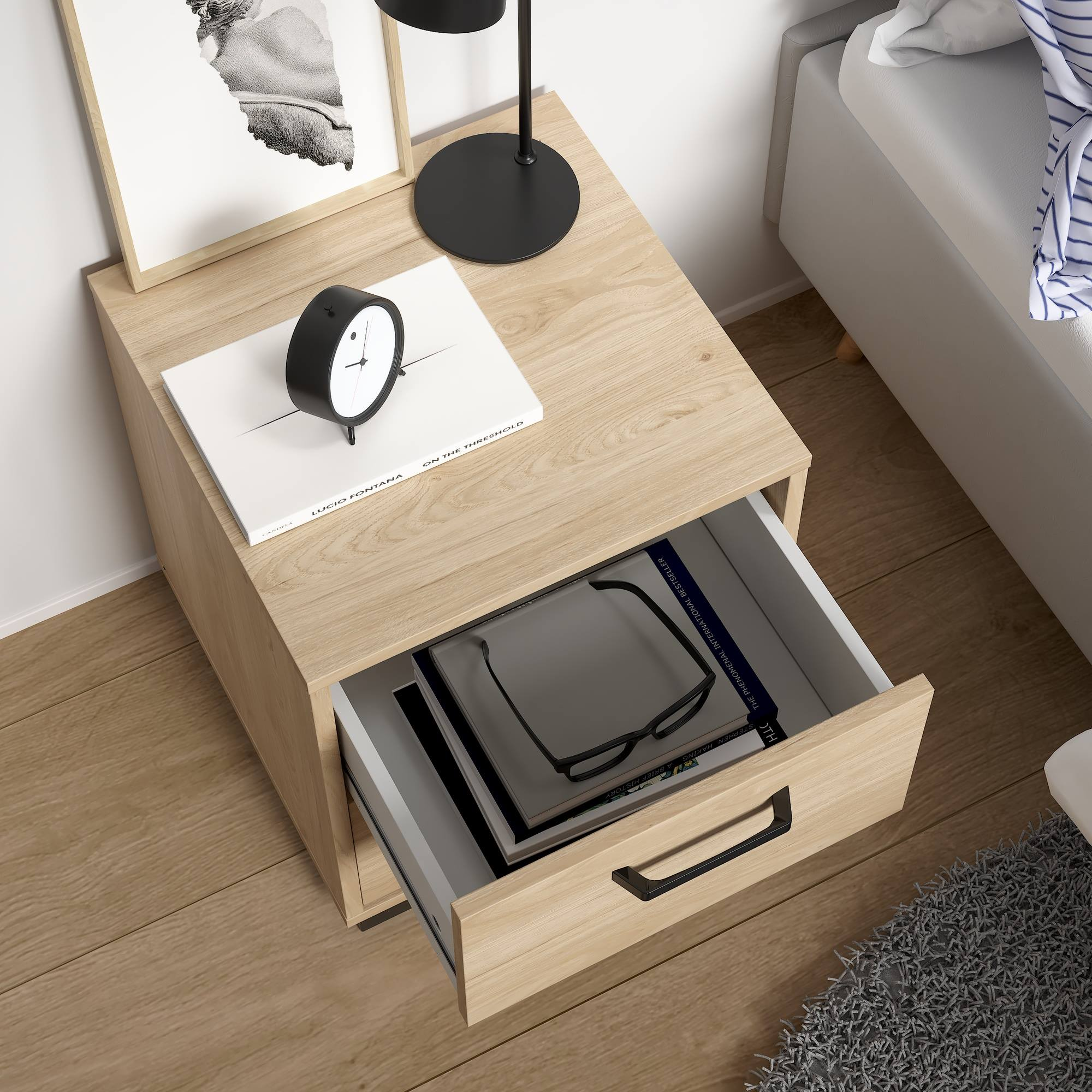 Una mesita de noche con un cajón abierto, libros, un reloj y gafas. Hay una cama a la derecha. Suelo de madera.